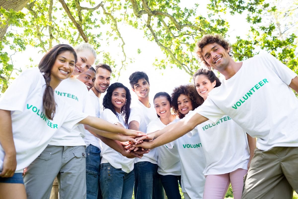 Group in white shirts stacking hands together outdoors under leafy trees