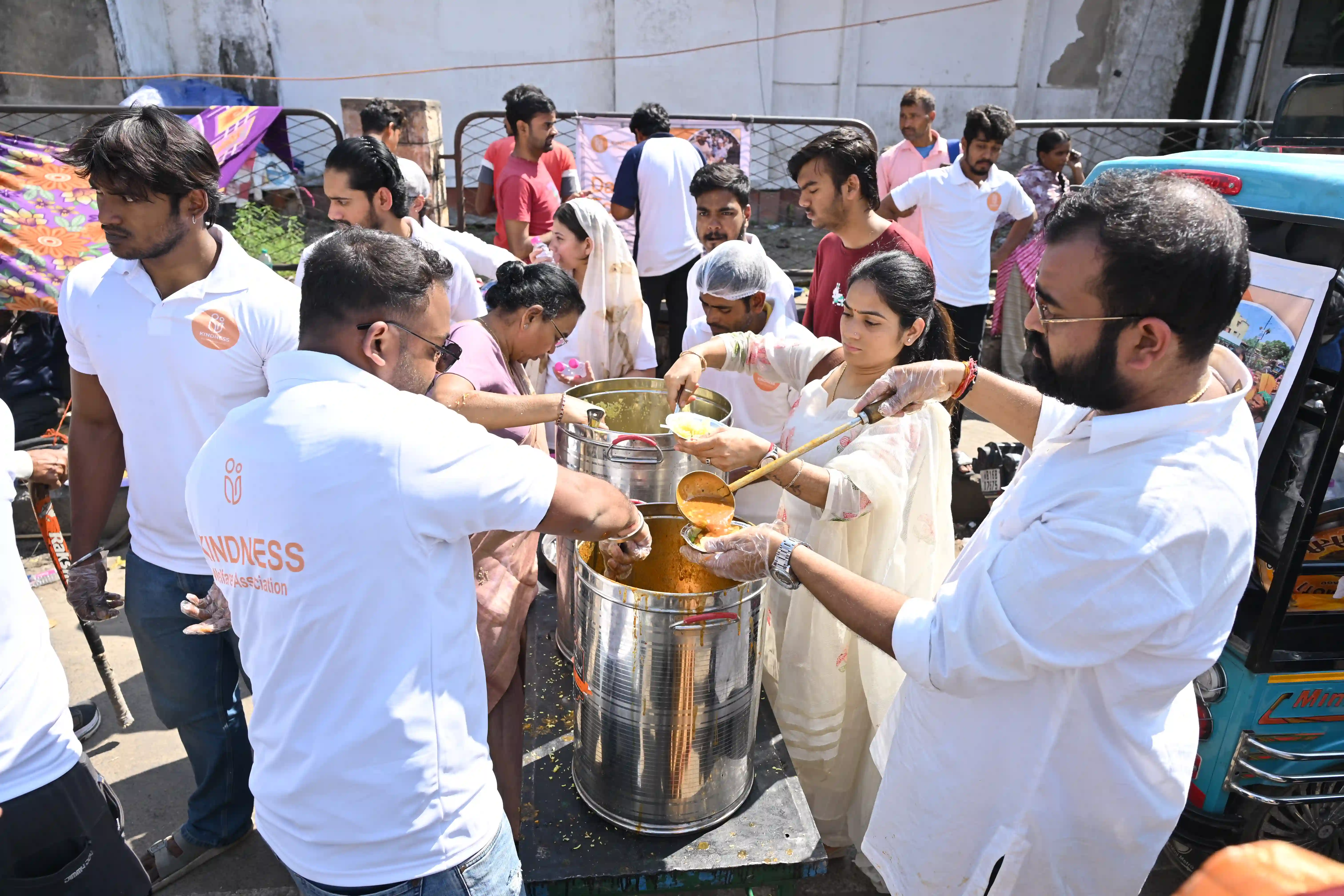 Volunteers serve drinks from large metal pots to a crowd on a sunny street.